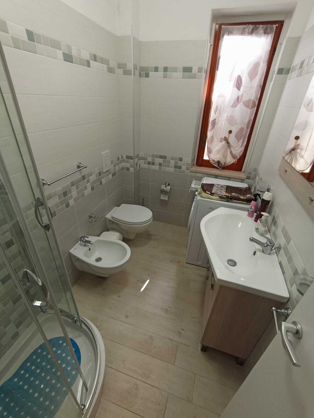 A bathroom with a shower, two toilets, a sink, and a window with a curtain.