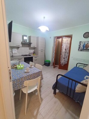 A compact room with a kitchenette, a dining table with chairs, a bed, and a single door.