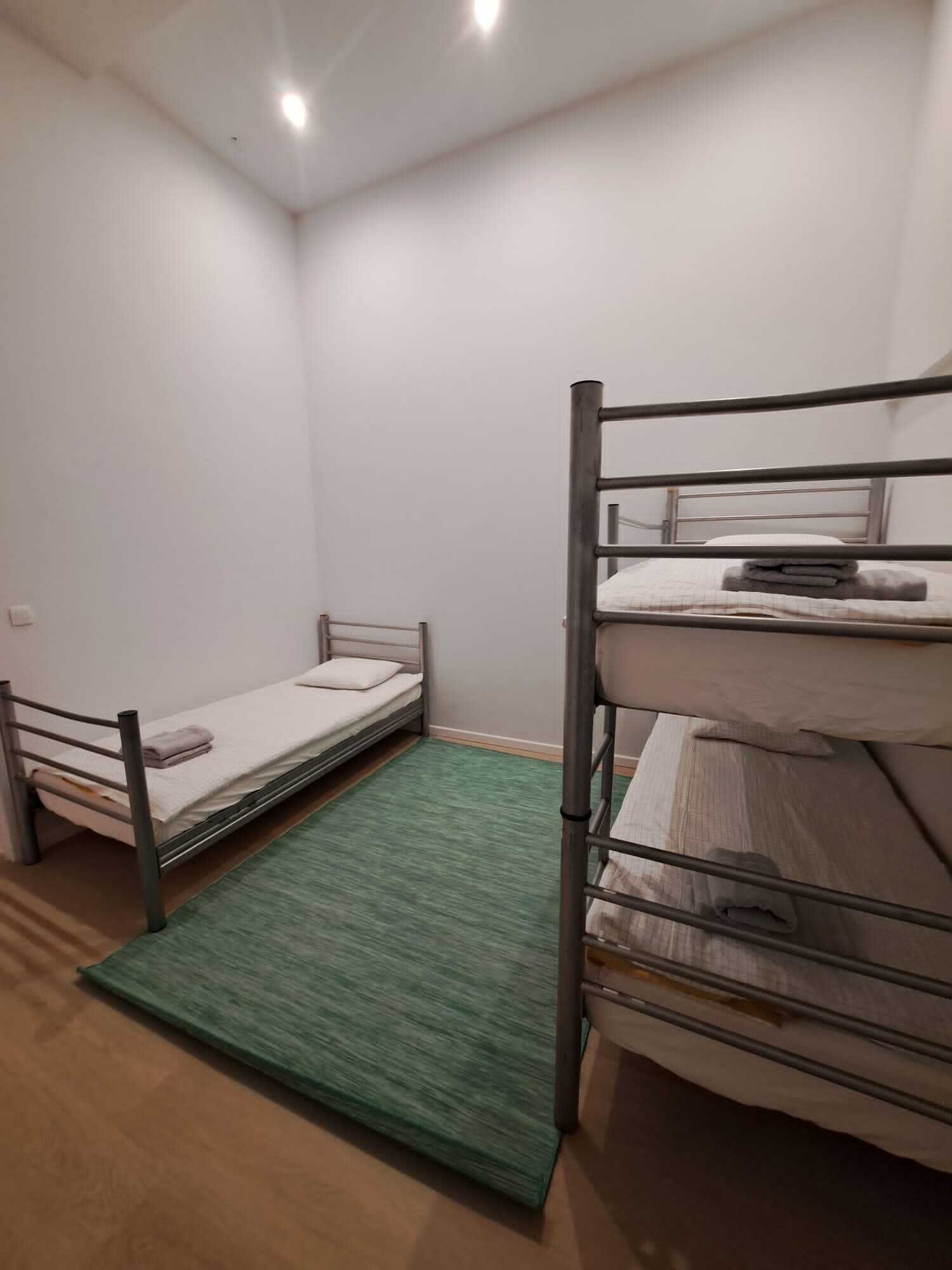 Un petit logement composé d’une seule pièce, avec deux lits superposés, un tapis vert et un mur blanc.