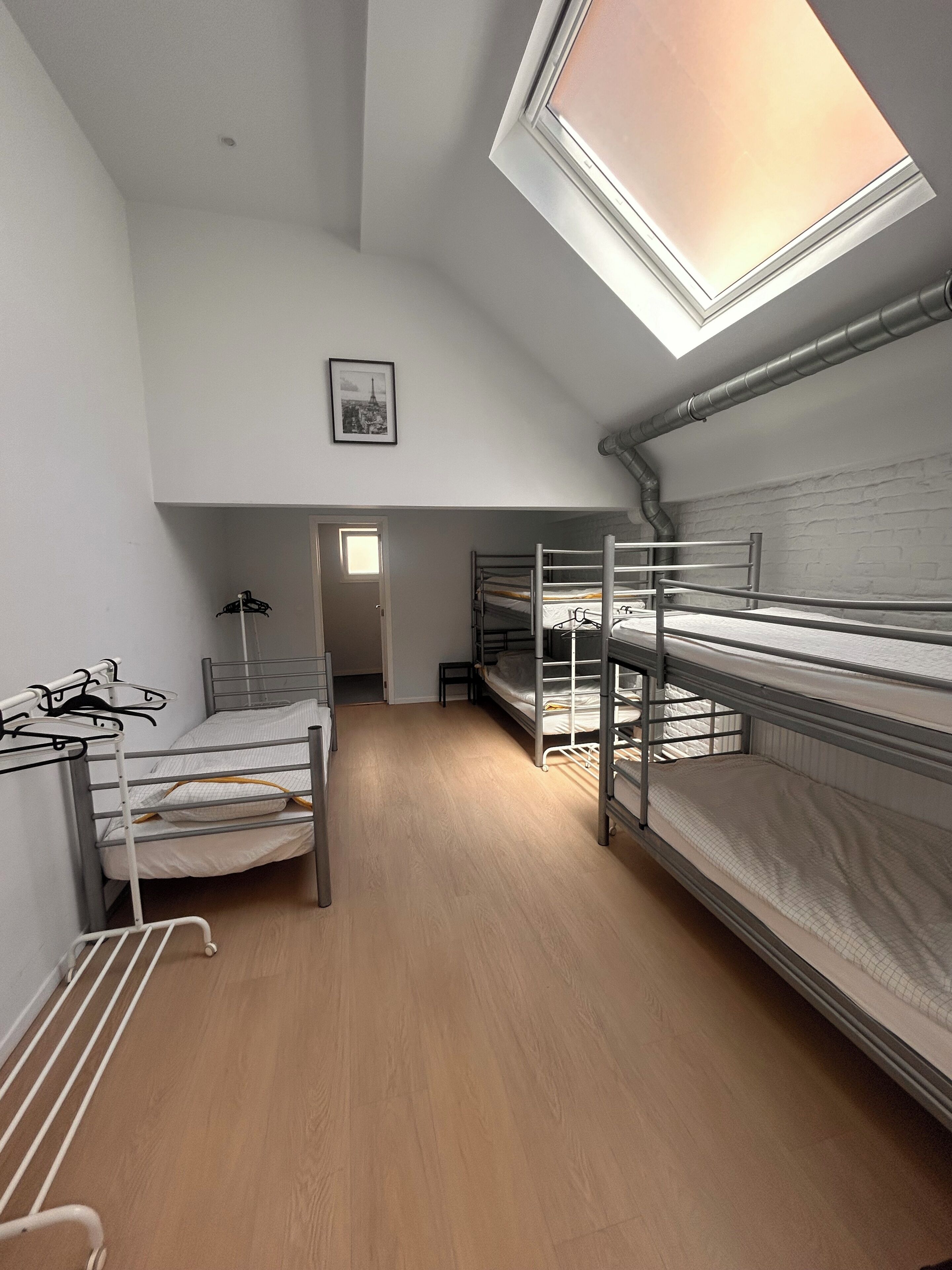 Een slaapzaal met stapelbedden, een dakraam, een lijst met een foto aan de muur, en een deur die naar een andere kamer leidt.