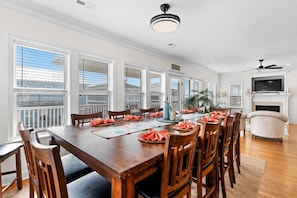 A dining room with a large wooden table set for six, surrounded by wooden chairs. There is a television mounted above a fireplace, and a ceiling fan is visible. - Second Row - Private Pools & Ocean Views by Williamson Realty (Ocean Isle Beach)
