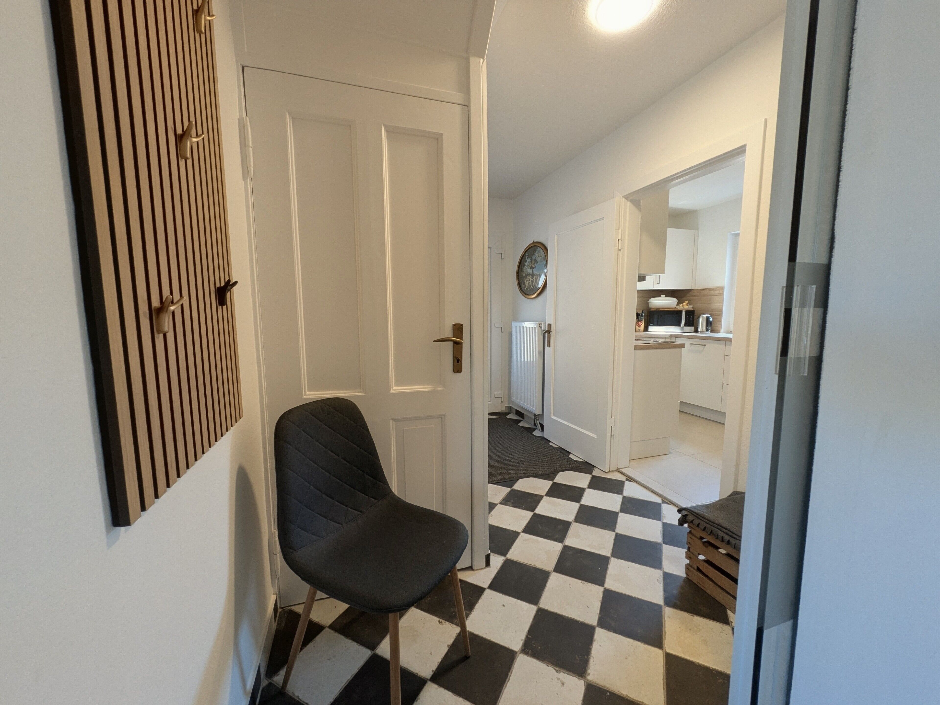 Een gang met een zwart-wit geruit vloerkleed, een stoel, een deur en een keuken die zichtbaar is door een open doorgang.