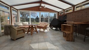 Uma sala de sol com teto de vidro, mesa de jantar, cadeiras, sofá e churrasqueira.