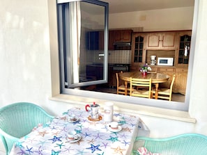 A dining area with a table set for two, a window with a view of a kitchen, and a wicker chair. - Adorable Flat With Balcony (Lignano Sabbiadoro)