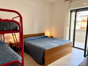 A room with a bunk bed, a single bed, a nightstand with a lamp, and a sliding glass door to a balcony. - Adorable Flat With Balcony (Lignano Sabbiadoro)