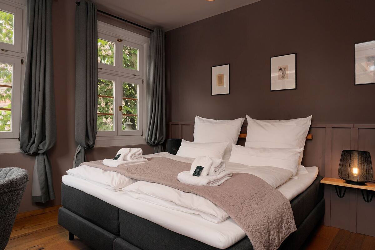 A neatly made bed with pillows and a blanket, a bedside table with a lamp, and a window with curtains.
