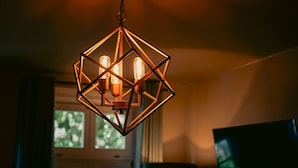 A geometric pendant light fixture with three bulbs, hanging in a room with a window and a television. - Wohnkultur DSIGNIO Digital Hotel (Ladenburg)