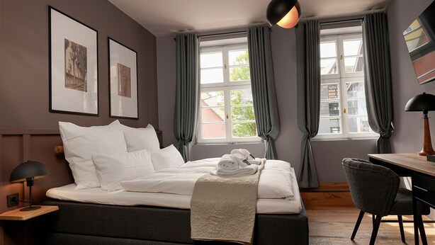 A bedroom with a bed, a desk, a chair, a lamp, and two windows with curtains. - Wohnkultur DSIGNIO Digital Hotel (Ladenburg)