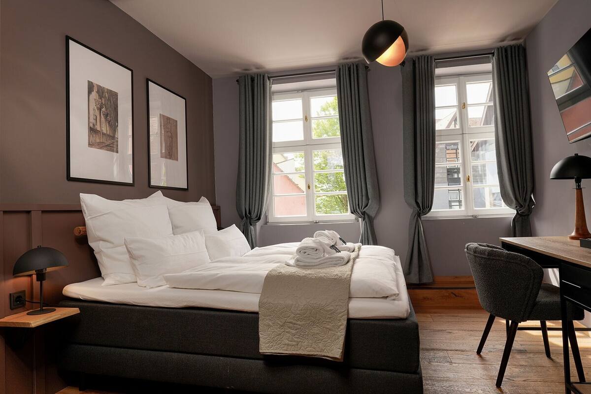 A bedroom with a bed, a desk, a chair, a lamp, and two windows with curtains.