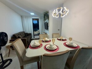 A dining area with a table set for six, a sofa, a TV, and a ceiling light fixture. - NOA - Miramar Hospedagens (Cabedelo)