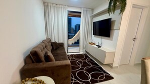 Un salon moderne avec un canapé marron, une table ronde blanche, un tapis à motifs, un téléviseur posé sur un meuble blanc et une vue sur les immeubles à travers une porte vitrée.