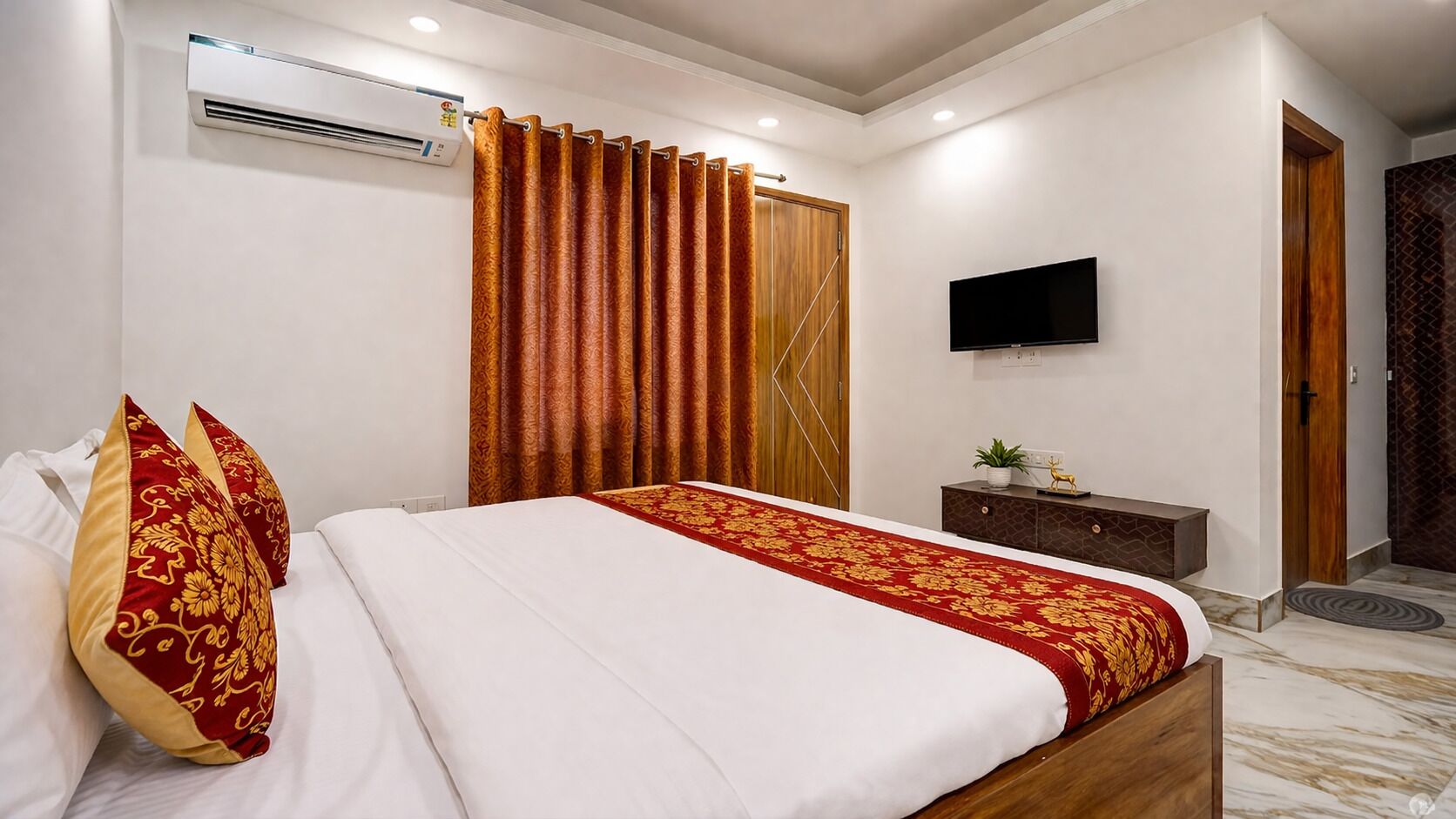 A hotel room with a bed, curtains, a flat-screen TV, a wooden door, and a small plant on a nightstand.