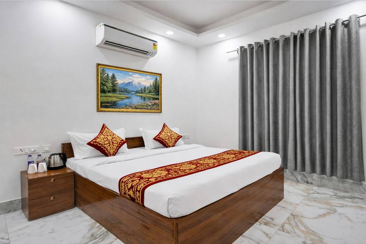 A hotel room with a bed, a nightstand, a painting, an air conditioner, and grey curtains.