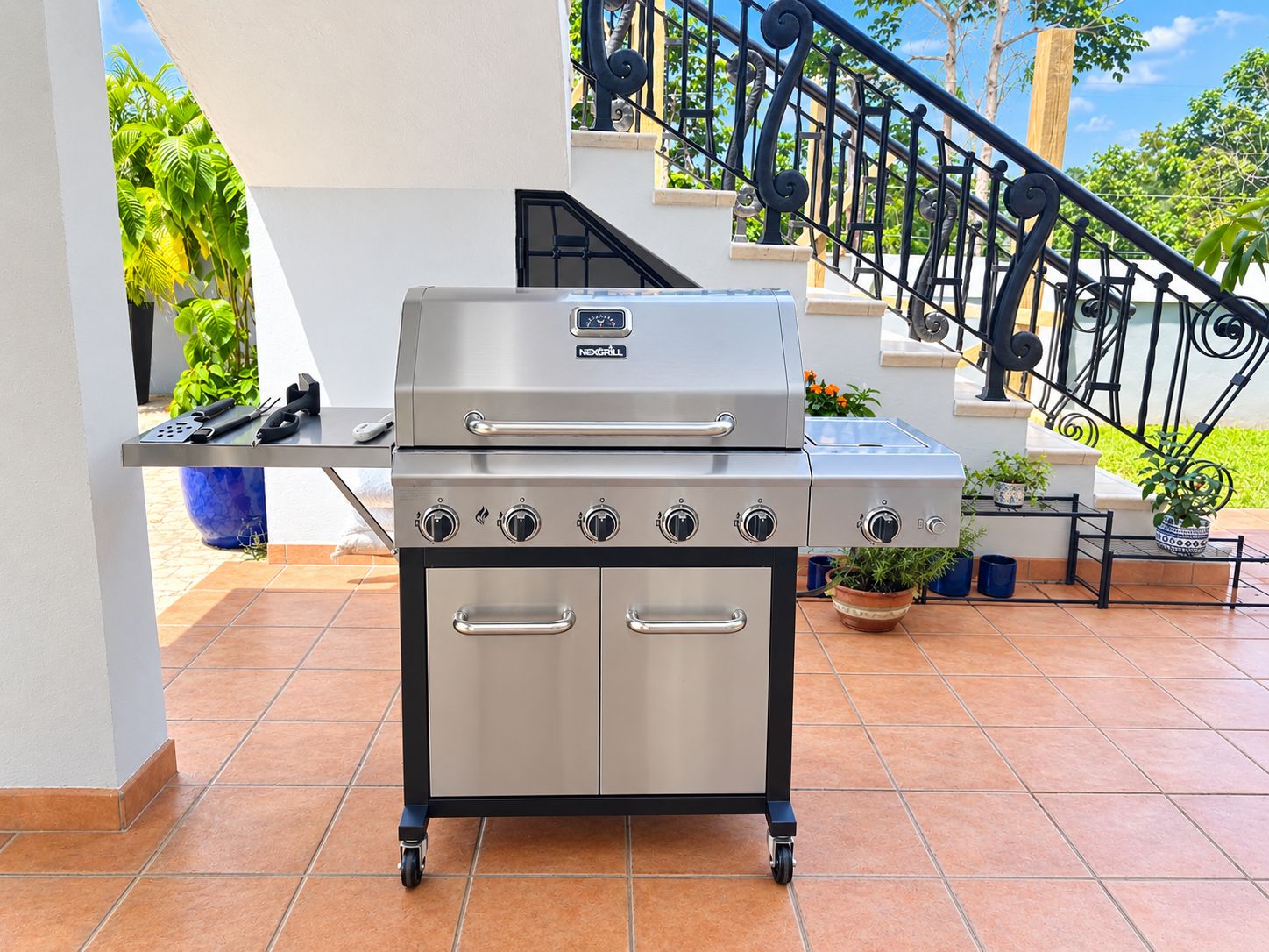 Un barbecue extérieur en acier inoxydable, doté d’un panneau de commande et d’un chariot, installé sur une terrasse carrelée, avec un escalier en arrière-plan.