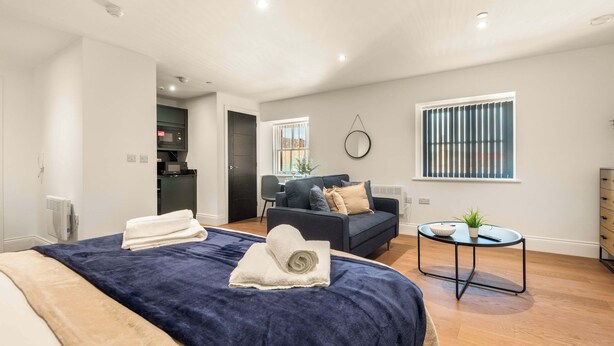 A modern bedroom with a large bed, a dark blue sofa, a small round table, and a window with blinds. - Contemporary Spacious Studio in Central Sheffield (Sheffield)