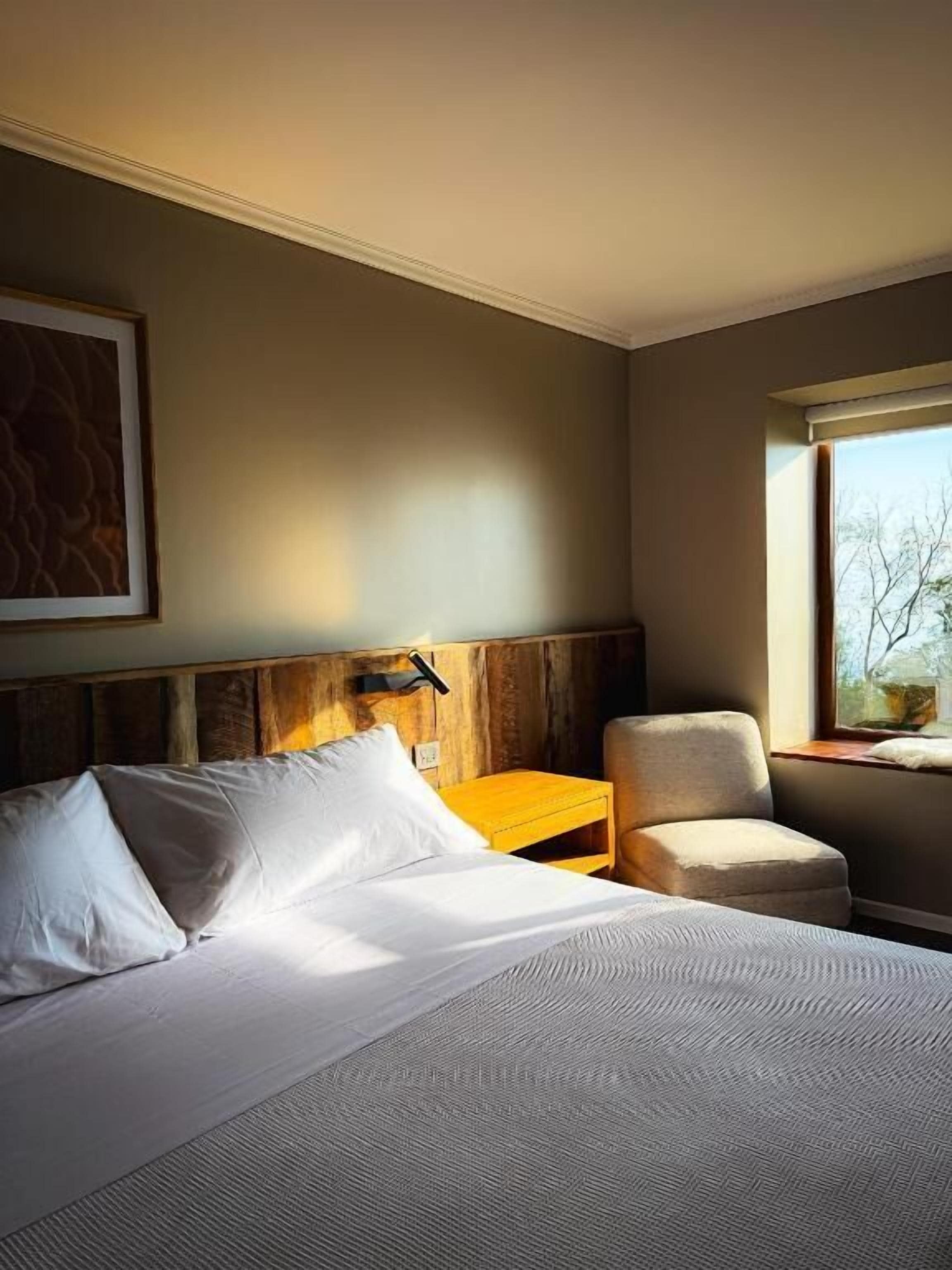 Una habitación de hotel con cama, una silla, una mesita de noche y un ventanal con vistas.