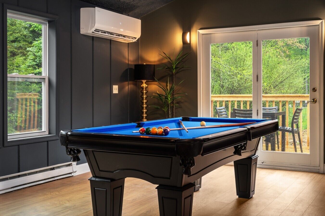 A pool table with balls, a lamp, and a potted plant in a room with a view through a glass door.