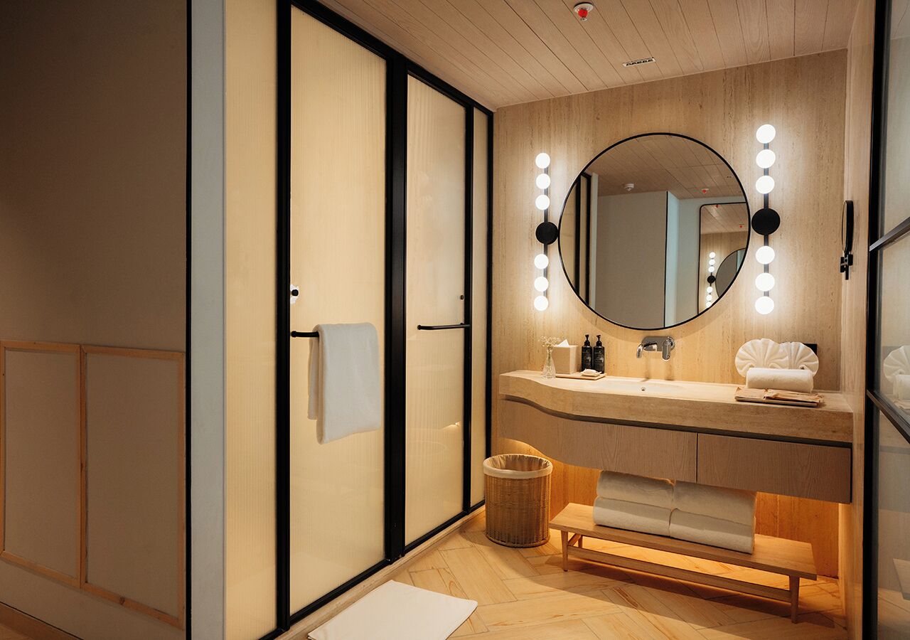 Un baño moderno con tocador de madera, un espejo redondo con iluminación, un banco y un cesto de basura.