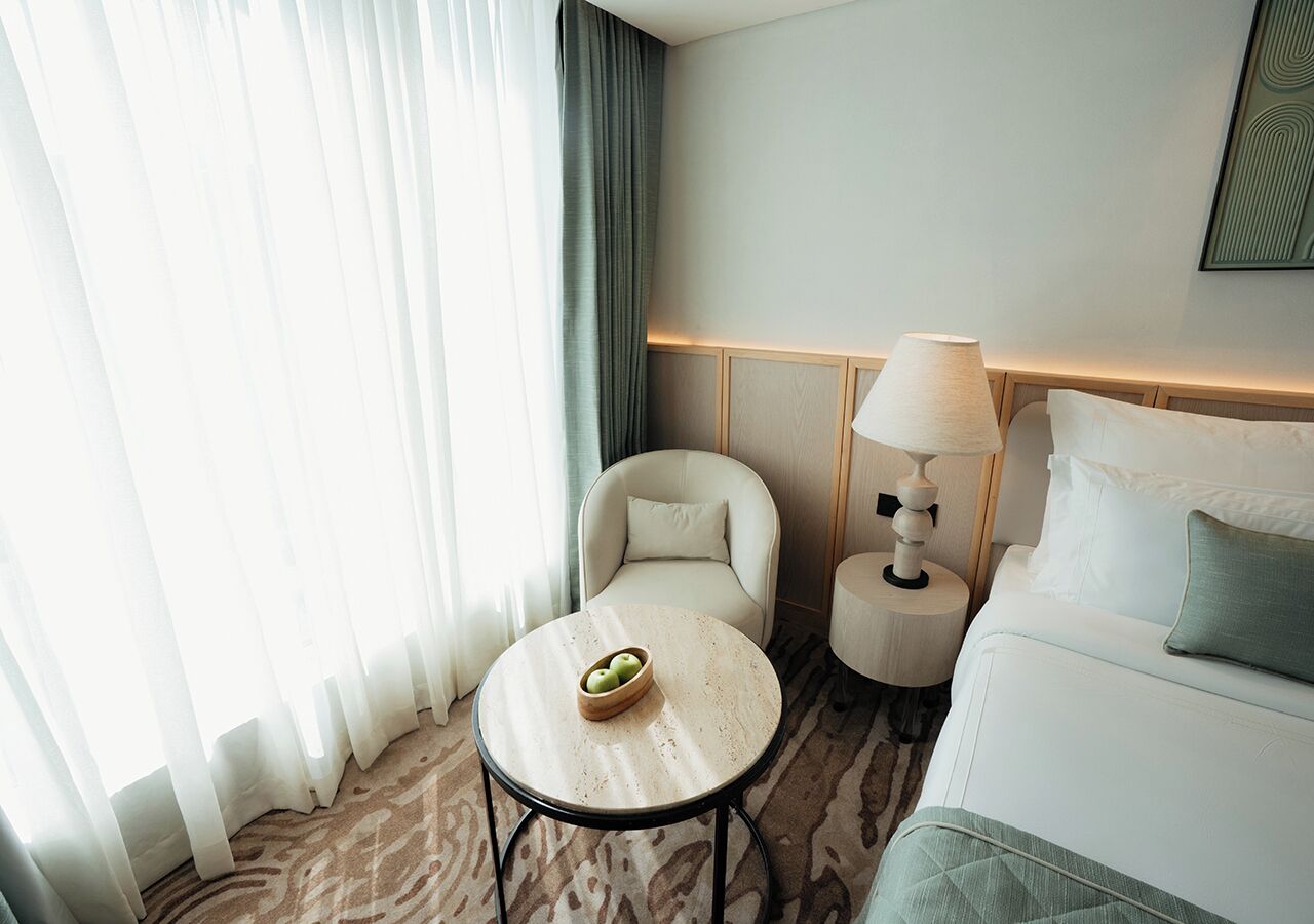 A hotel room with a bed, a round table with a bowl of fruit, a chair, a lamp, and a view of the outdoors.