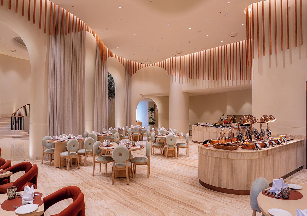 A spacious hotel dining area with multiple round tables and chairs, a central bar, and a staircase leading to an upper level.