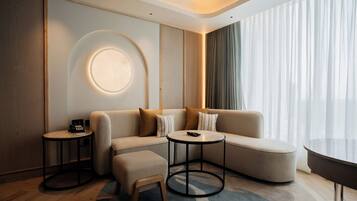 A modern hotel room with a sofa, ottoman, two round tables, a small side table, and a large window with curtains.