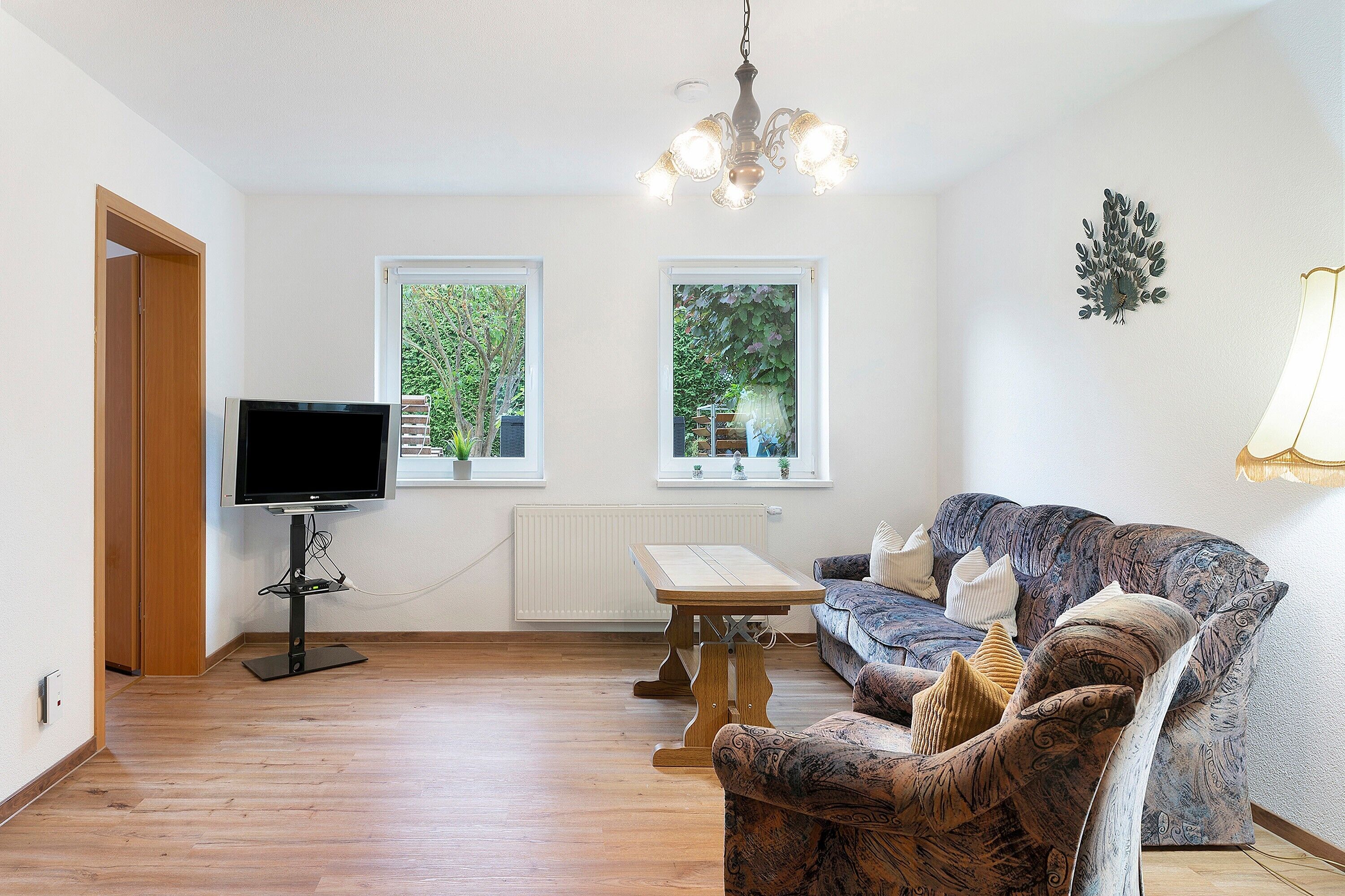 Ein Wohnzimmer mit Fernseher, Tisch, Sofa und zwei Fenstern mit Blick auf Grünflächen.