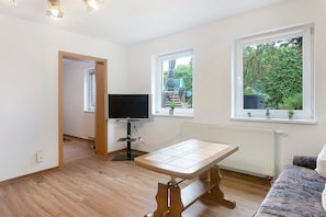 Ein Zimmer mit Holzboden, Fernseher, Tisch und zwei Fenstern mit Blick auf Grünflächen.