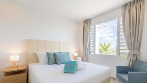 Quarto de hotel com cama, mesa de cabeceira, cadeira, janela com cortinas e vista para palmeiras.