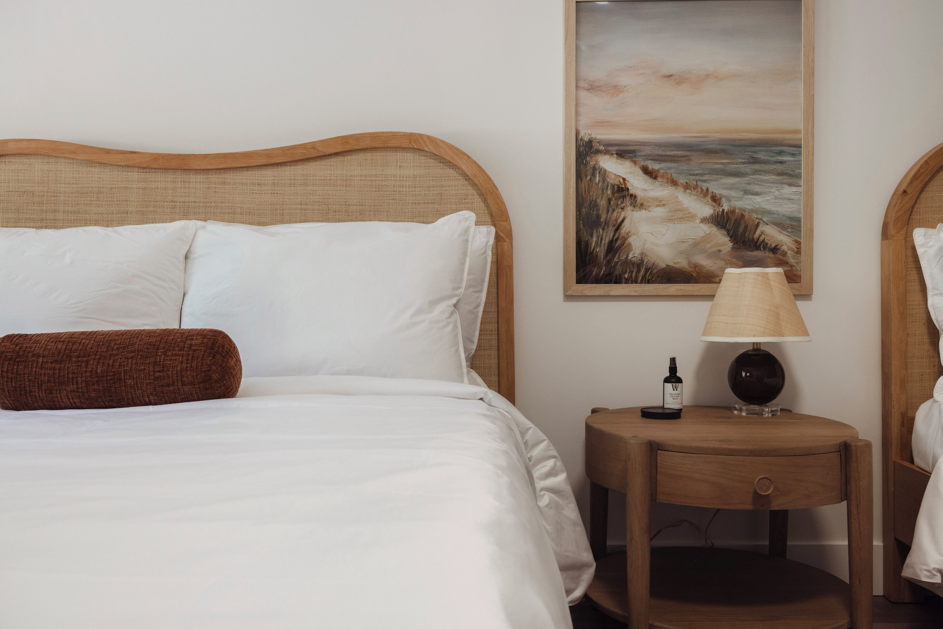 Una cama bien hecha con sábanas blancas y un almohadón marrón, una mesita de noche de madera con una lámpara y una botella, y un cuadro de un paisaje costero en la pared.