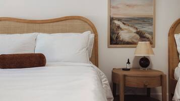 Una cama bien hecha con sábanas blancas y un almohadón marrón, una mesita de noche de madera con una lámpara y una botella, y un cuadro de un paisaje costero en la pared.