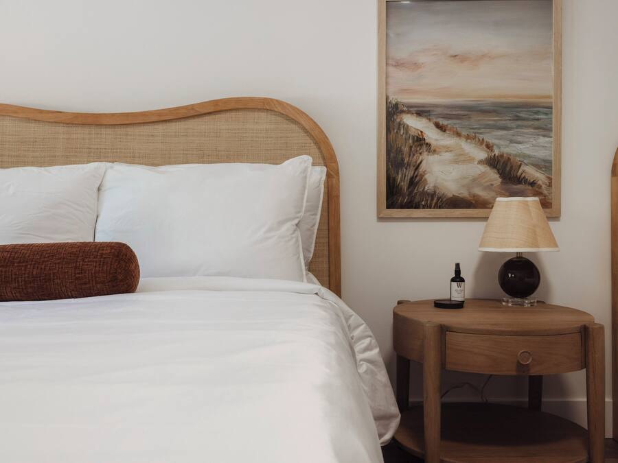 Una cama bien hecha con sábanas blancas y un almohadón marrón, una mesita de noche de madera con una lámpara y una botella, y un cuadro de un paisaje costero en la pared.