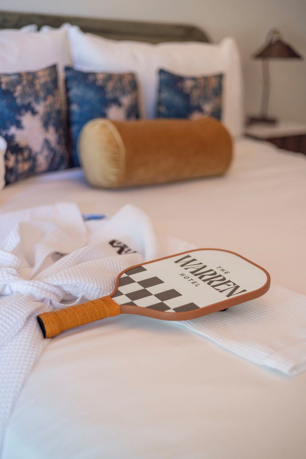 A hotel bed with a checkered paddle, a rolled towel, and a pillow with a blue pattern.