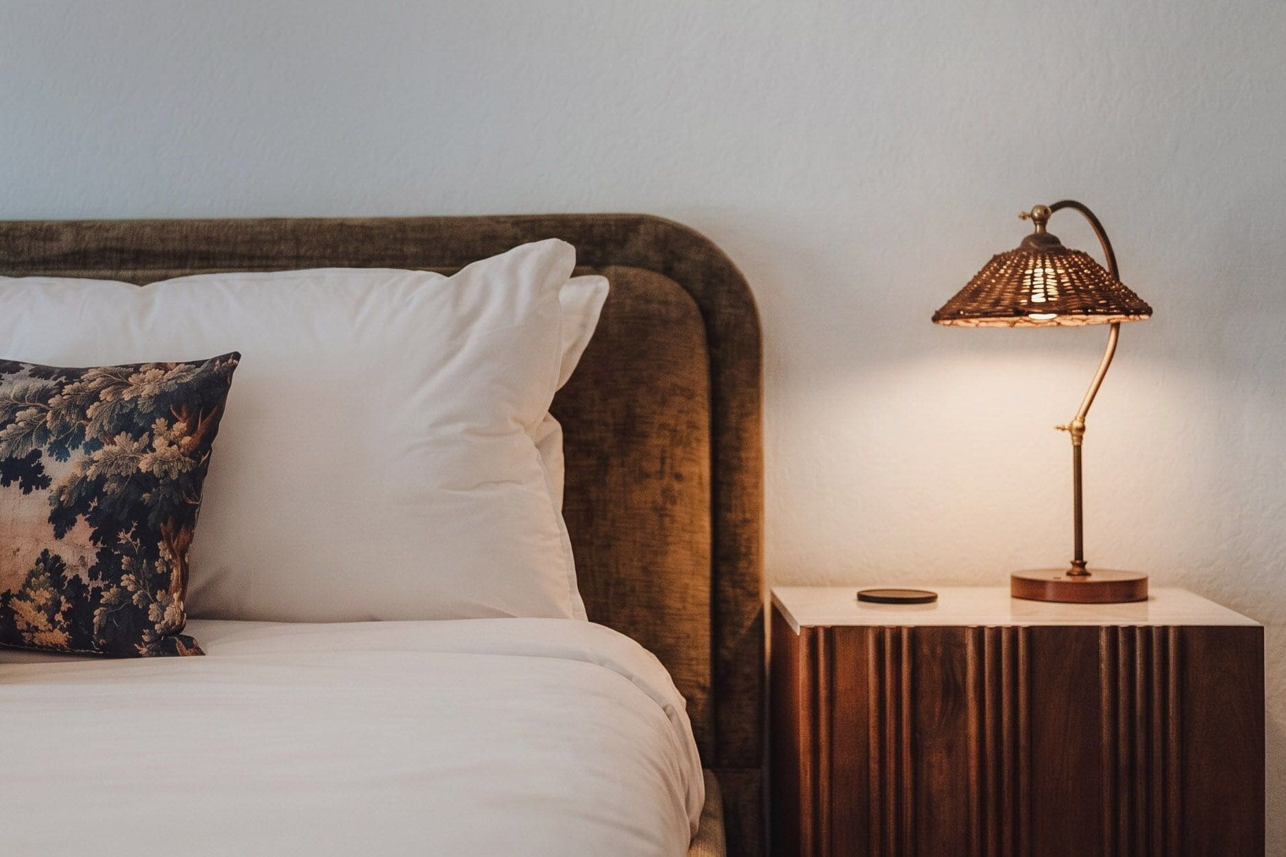 A bed with a white pillow and a decorative pillow, a bedside table with a lamp, and a wooden nightstand.
