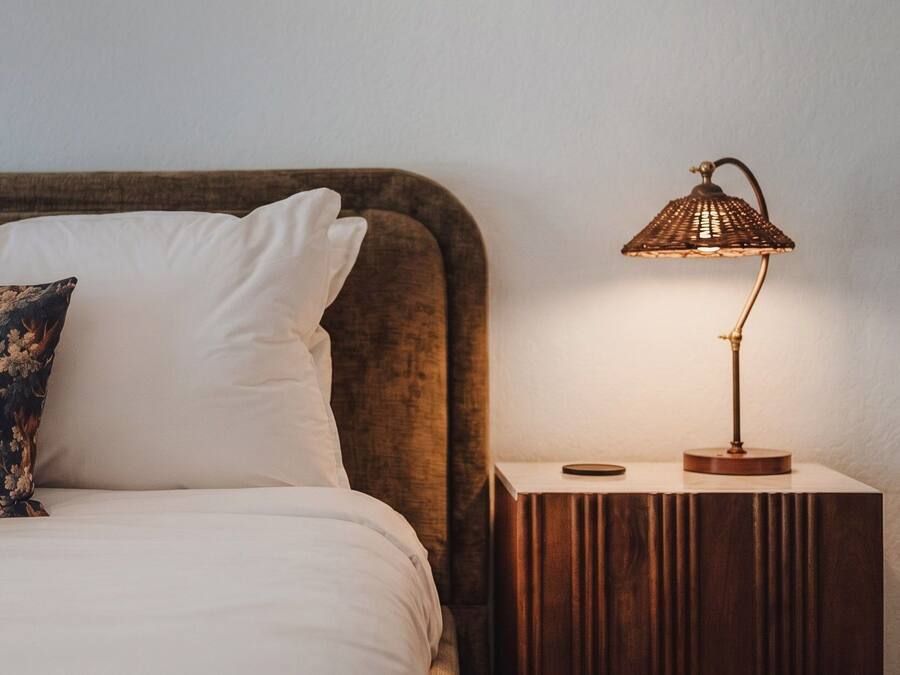 Una cama con un almohadón blanco y un cojín decorativo, una mesita de noche con una lámpara y una mesita auxiliar de madera.