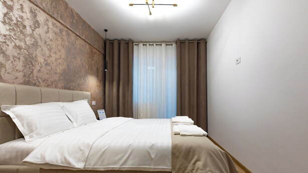 A bedroom with a bed, curtains, a wall with a textured pattern, and a ceiling light fixture. - Cozy & Modern Apartment Near Arena Mall, Free Parking, Wi-Fi ,Ac. (Bacau)