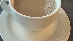 A white cup of coffee on a saucer with a textured surface, a white mug, a small plate with a condiment jar, and a silver teapot on a table.