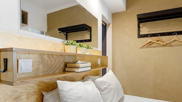 A hotel room with a wooden headboard, a bedside table with a mirror, a shelf with books and plants, a hanging coat rack, and a bed with white pillows.