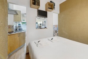Una habitación con una cama, un televisor montado en la pared, una zona de cocina y un espejo que refleja el cuarto.