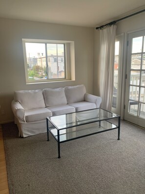 Una sala de estar con un sofá blanco, una mesa de centro de vidrio, una ventana con cortinas y vistas a edificios.