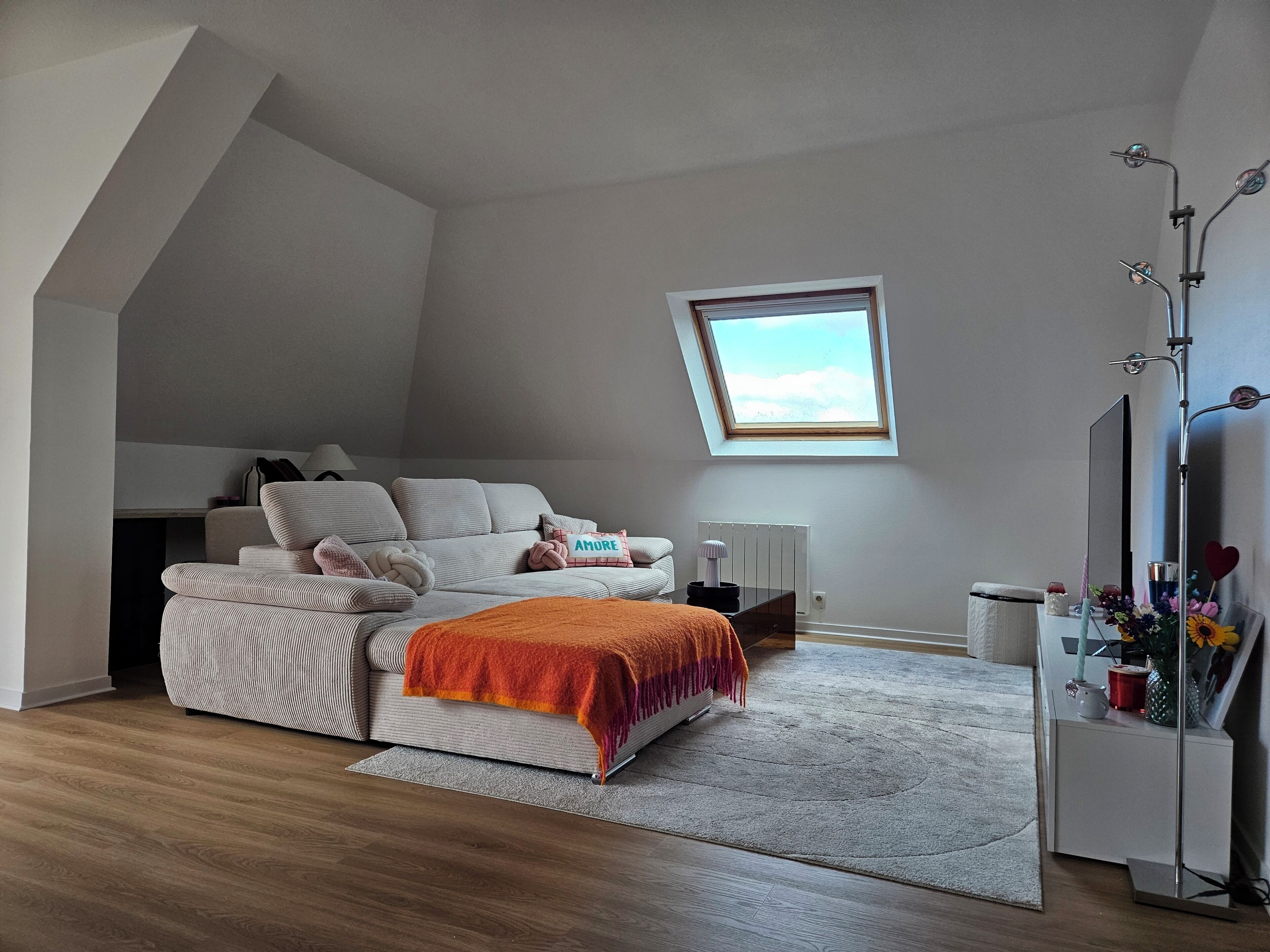 A loft-style bedroom with a sloped ceiling, a large window, a sofa, a bed, a radiator, and a bedside table with a mirror.