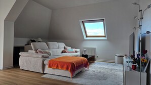 Um quarto estilo loft com teto inclinado, uma janela grande, um sofá, uma cama, um radiador e uma mesa de cabeceira com um espelho.