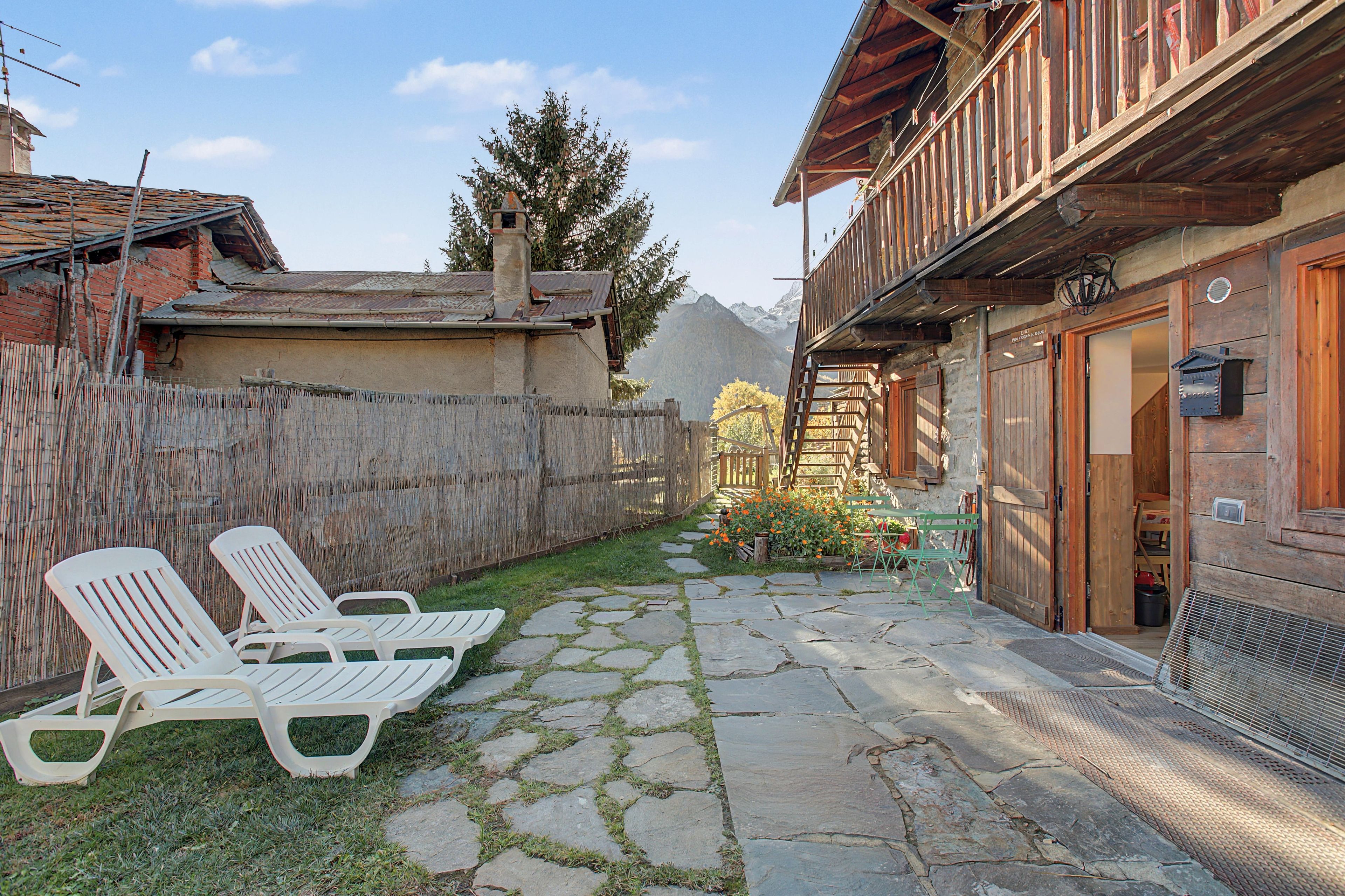 A stone-paved courtyard with two white lounge chairs, a wooden fence, and a mountain backdrop.
