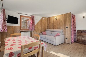 Une pièce chaleureuse avec un mur en bois, un canapé gris, une table à manger recouverte d’une nappe à motifs et un téléviseur fixé au mur.