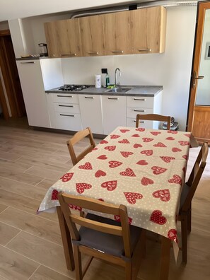 Une cuisine avec des meubles blancs, une cuisinière à gaz et un évier. On y trouve une table à manger recouverte d’une nappe à motifs en forme de cœur et des chaises en bois.
