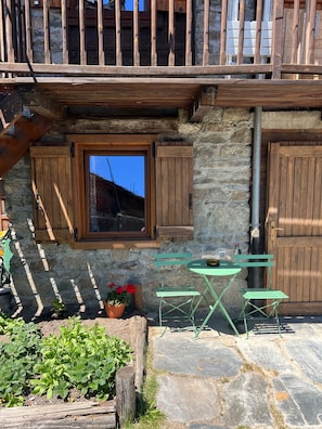 Un bâtiment rustique en pierre, doté d’un balcon en bois, d’une fenêtre avec des volets et d’un petit espace extérieur aménagé avec une table et des chaises.