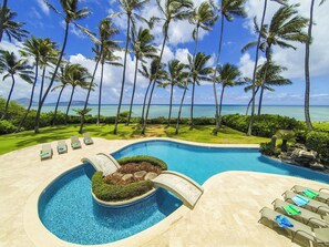 A pool with a curved design, surrounded by lounge chairs and palm trees, with a view of the ocean.