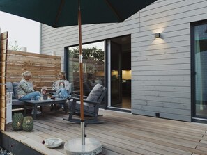 Un espace extérieur aménagé avec une terrasse en bois, une partie couverte et un bâtiment moderne doté de grandes fenêtres.