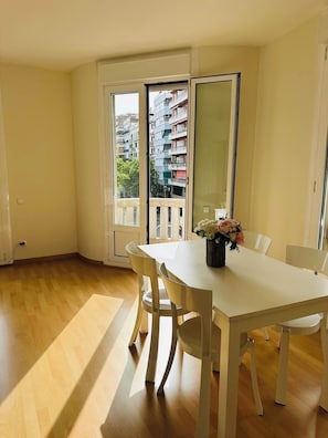 Une pièce avec une table et des chaises blanches, un vase rempli de fleurs et une vue sur un bâtiment, visible à travers une porte vitrée.