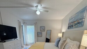 Ein Schlafzimmer mit Bett, Fernseher, Deckenventilator, Nachttisch, Lampe, einem Fenster mit Vorhängen und einem Bild an der Wand.