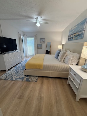 A bedroom with a bed, a TV, a ceiling fan, a nightstand, a lamp, a window with curtains, and a painting on the wall. - Newly renovated 2-bedroom, 2-bathroom condo located in sunny Sarasota, Florida. (Sarasota)